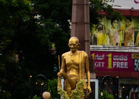 A golden statue of Gandhi set in an urban environment with surrounding greenery and architecture.