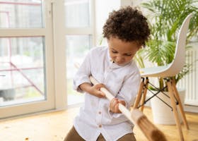 Young child with curly hair playfully sweeping a modern indoor space with a broom.
