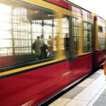 A woman in a yellow coat waits as a red train passes in a bustling station.