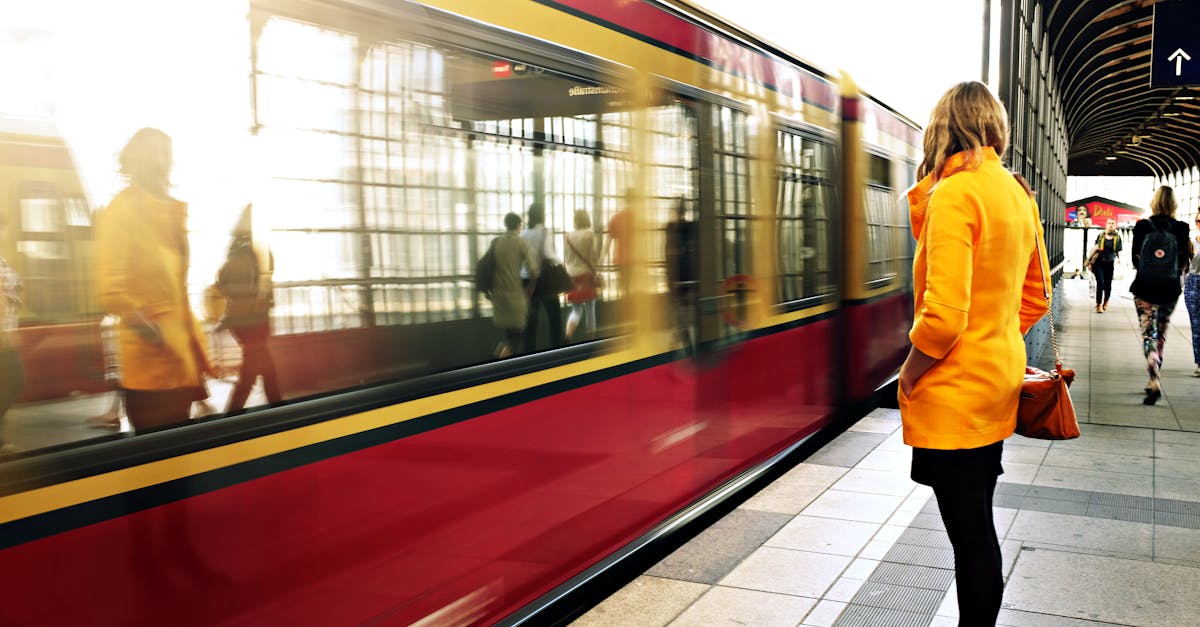 A woman in a yellow coat waits as a red train passes in a bustling station.